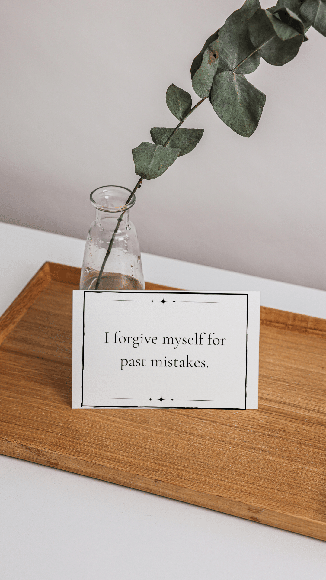 Mantra card that reads "I forgive myself for past mistakes" sits propped against a clear vase with a branch of eucalyptus in it.